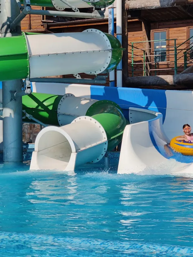 Closeup of green and white waterslides at Thermal Aqua Park Elbrus with child sliding on a yellow raft into the blue pool water.