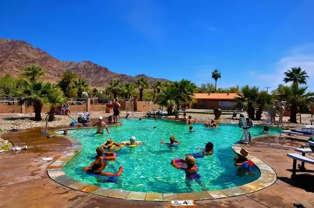 People relaxing on flotation rings in a clear turquoise hot spring pool surrounded by palm trees and desert mountains at Glamis North Hot S.