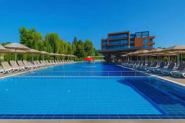 Large outdoor pool at Regnum Banya Thermal Pools with multiple sun umbrellas and long rows of lounge chairs under a bright blue sky in Bany.