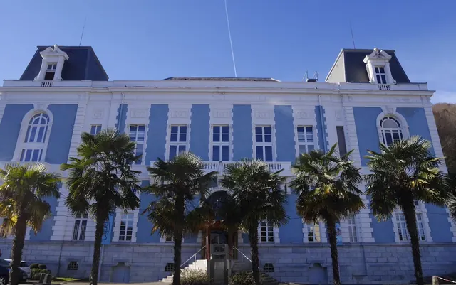 Blue and white historical building with mansard roofs and palm trees in front, located near the Aquensis Spa Thermal in Bagneres-de-Bigorre.