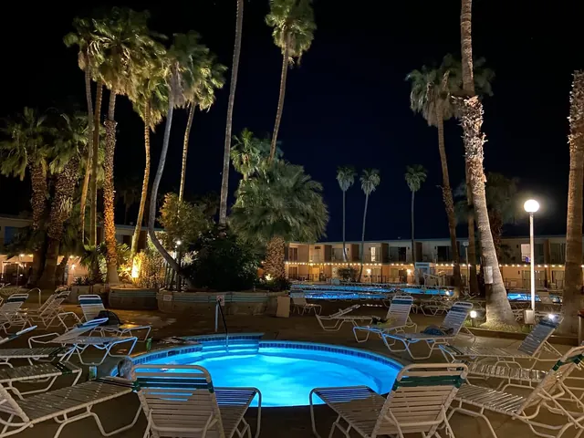 Night view of illuminated blue hot spring soaking pool at Desert Hot Springs Spa Hotel with empty lounge chairs and palm trees under dark s.