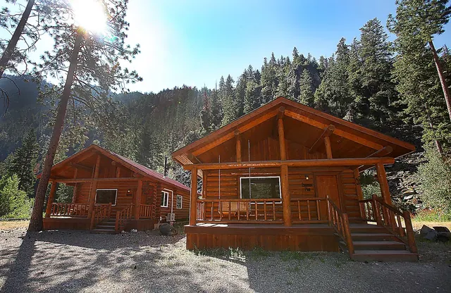 Two wooden log cabins with porches sit under bright sunlight near a forested mountain at Quinn's Hot Springs Resort, Montana.