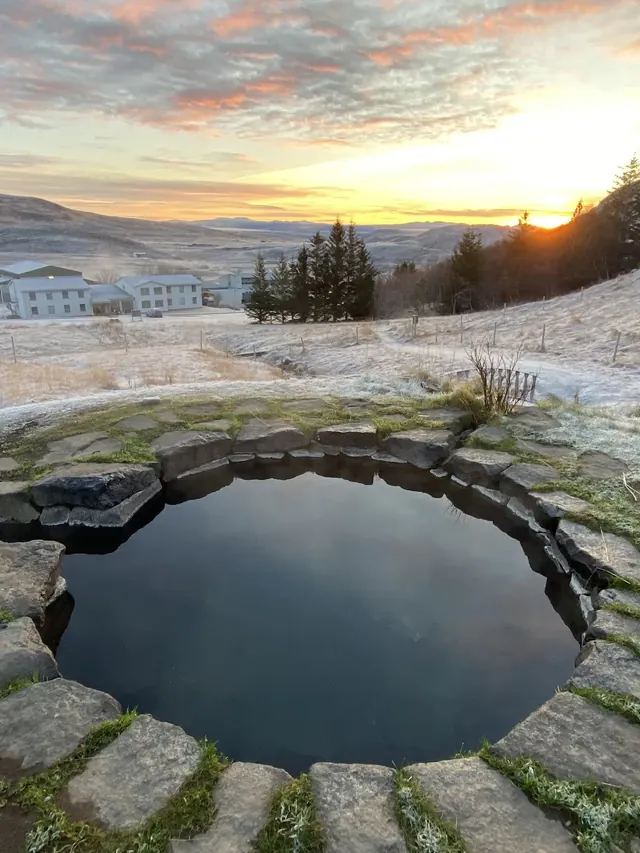 Gudrunarlaug hot spring pool steaming gently at sunset with frosted grass and buildings in the distance in Budardalur Iceland.