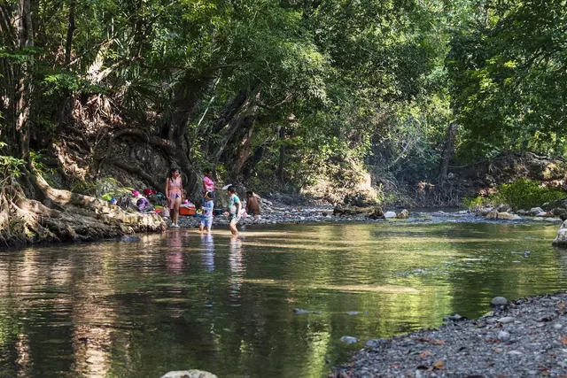 Children and adults wading and standing in a shallow creek surrounded by trees and rocks in a natural setting near Finca El Paraiso Hot Spr.