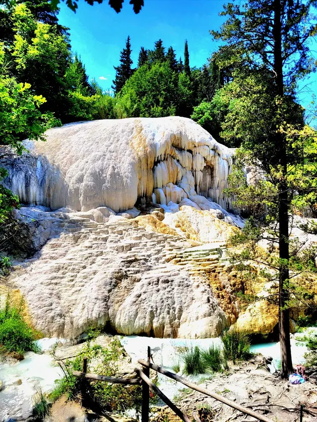 White calcium deposits form a rounded natural hot spring formation surrounded by green trees under bright skies.