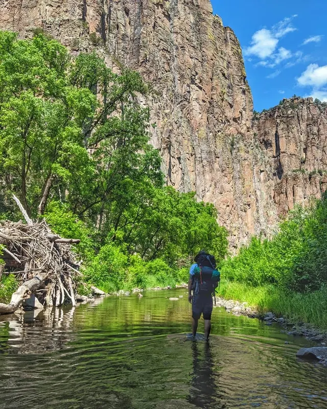 A person with a large backpack wades through a shallow stream with tall cliffs and green trees in the background under a blue sky.