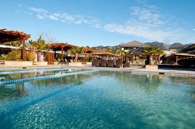 Clear blue outdoor mineral pool at Calistoga Spa Hot Springs with lounge chairs and wooden pergolas under a partly cloudy sky.