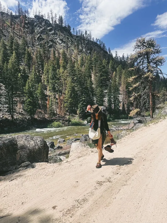 Woman hiking on a dirt trail beside a river with pine-covered hills in the background near Sacajawea Hot Springs, Lowman, Idaho, USA