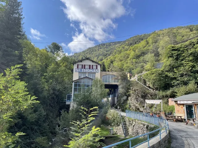 Les Bains de Saint-Thomas hot spring building nestled on a hillside with dense trees and a cloudy blue sky near Fontpédrouse, France.