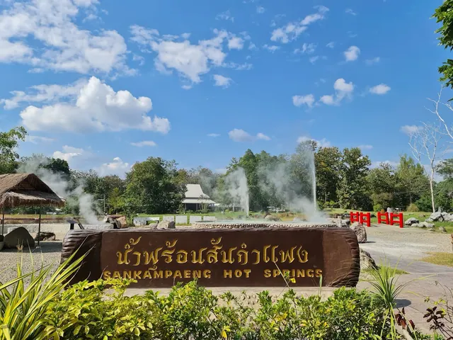 Wooden entrance sign for San Kamphaeng Hot Springs with steaming geysers and trees on a sunny day near Chiang Mai, Thailand.