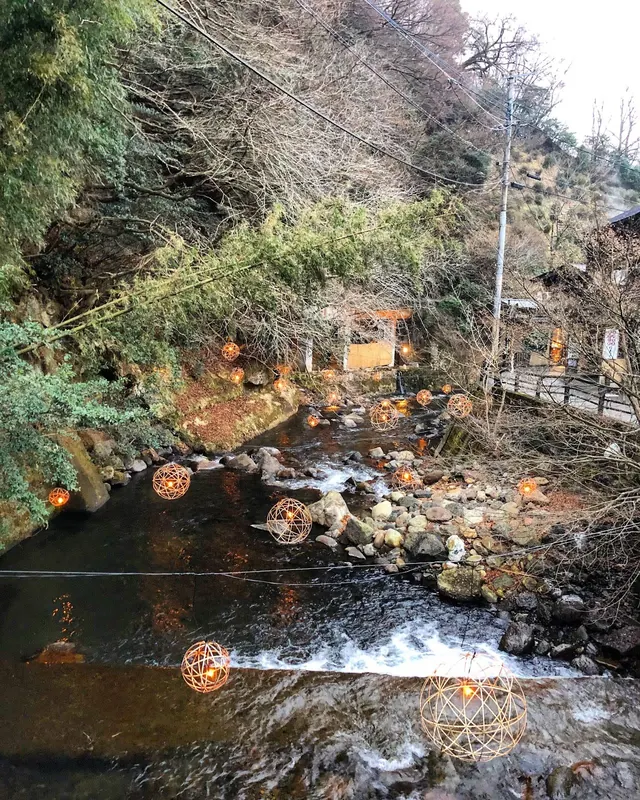 Stream with spherical wire lanterns floating above the water and bare trees surrounding it in Kurokawa Onsen.