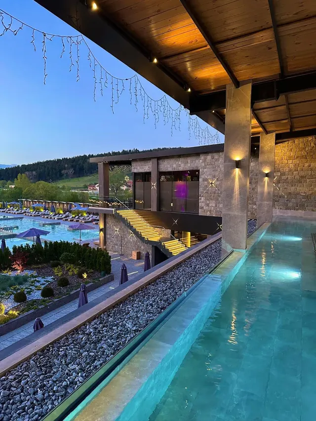Evening view of Pulse Therme Banya outdoor pool and stone building with illuminated stairs and purple umbrellas near the forest in Banya, B.