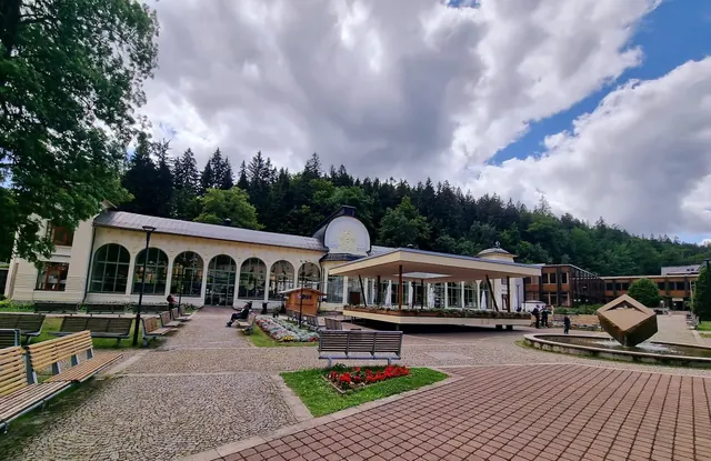 Public park area with benches, flower beds, and a white pavilion in front of a large historic building surrounded by forest.