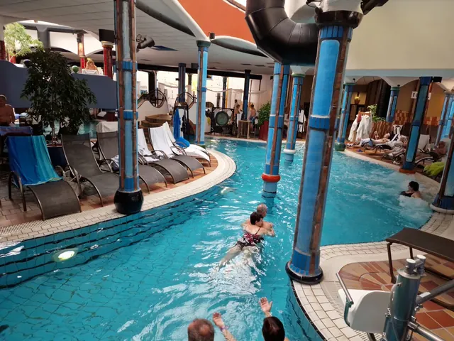 Indoor hot spring pool surrounded by tall blue columns and lounge chairs with several people relaxing in the water at Rogner Bad Blumau Aus.