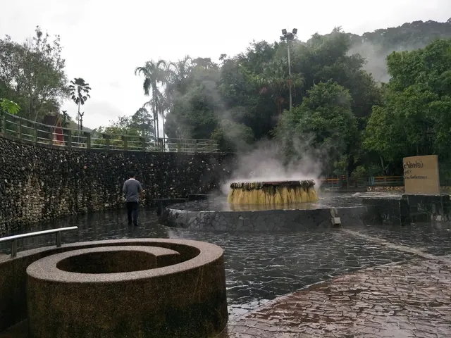 Wide view of the Raksawarin Hot Springs pool area showing a man walking near steaming mineral pools bordered by stone walls and vegetation.