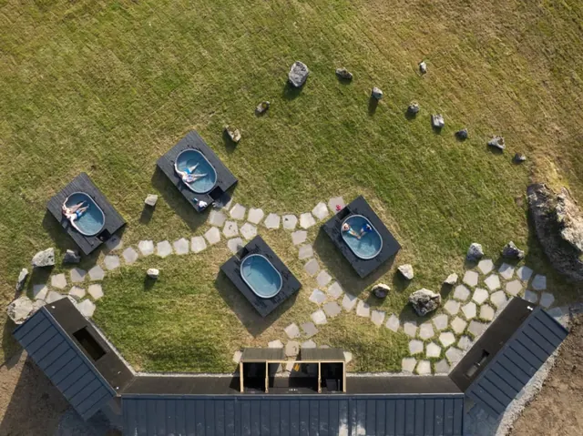 Aerial view of five black-framed tubs with clear water set on a stone path in green grass at Hoffell Hot Tubs Iceland.