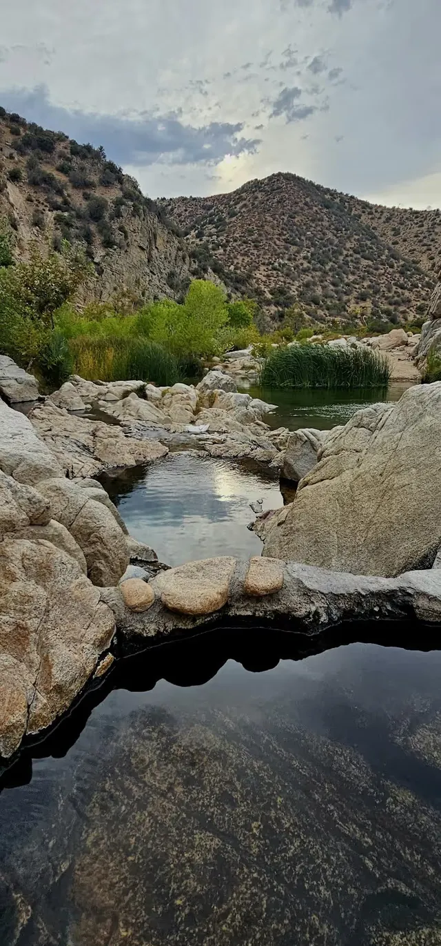 Natural hot spring pool surrounded by large rocks and green bushes with hills in the background at Deep Creek Hot Springs, California.