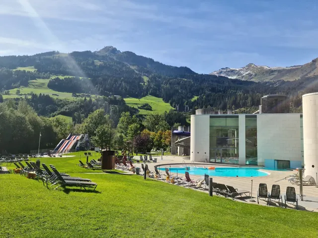 Lounge chairs and a clear outdoor pool with mountain hills and partly cloudy sky near Alpentherme Gastein in Bad Hofgastein, Austria.