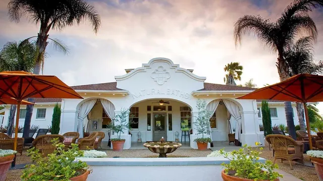 White building labeled Natural Baths with courtyard seating, umbrellas, and palm trees under a colorful sky in Calistoga, California.