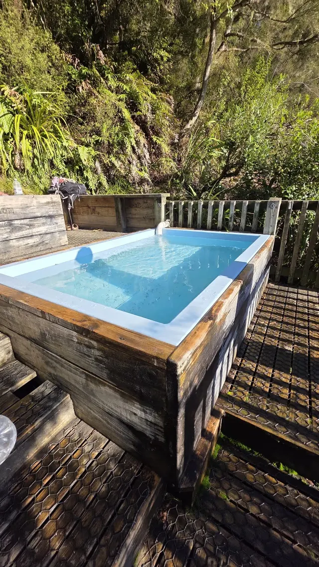 Clear water filled rectangular wooden pool surrounded by lush plants on a sunny day at Mangatutu Hot Springs.