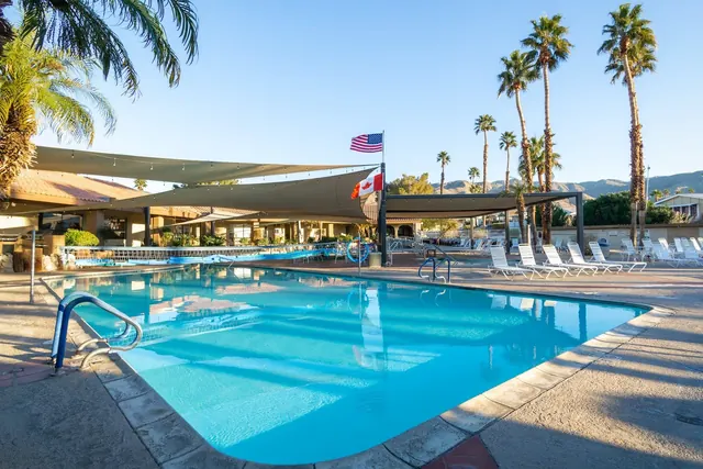 Wide rectangular outdoor pool at Caliente Springs Resort in Desert Hot Springs California with white lounge chairs and two flags on a pole.