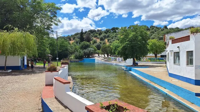 Outdoor soaking pool with a few people near white buildings painted with blue trim, set against a background of green hills at Termas da Su.