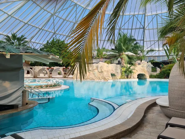 Clear blue water of an indoor mineral pool at Therme Erding, Germany with rock formations and tropical plants visible under glass roof.