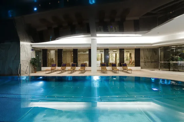 Indoor hot spring pool with lit blue tiles and woven lounge chairs aligned against a marble wall inside Hotel Thermal Pool in Karlovy Vary.