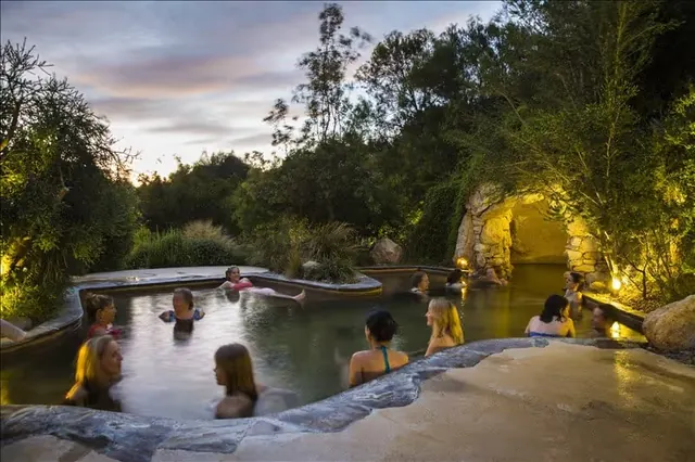 Group of people relaxing in an outdoor hot spring pool near a small stone cave entrance at Peninsula Hot Springs, Victoria, Australia at du.