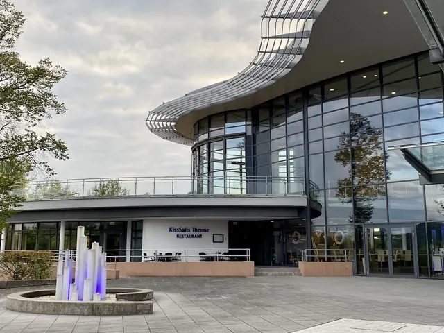 Facade of KissSalis Therme restaurant with tall glass windows and modern roof design next to stone plaza and decorative water feature in Ba.
