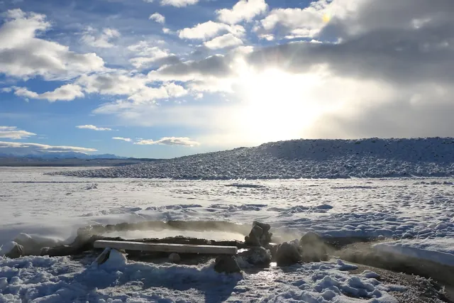 Steaming outdoor hot spring pool surrounded by snow under a cloudy sky with the sun peeking through near Mammoth Lakes California.