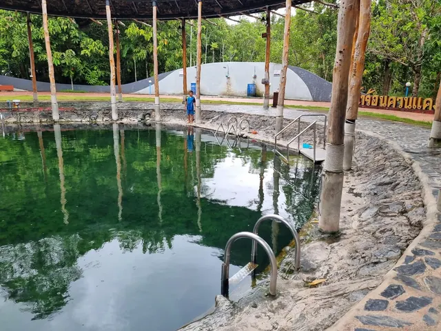 Calm green water fills a round hot spring pool shaded by a wooden pavilion with natural stone seating in Kantang Hot Spring Forest Park.