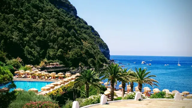 Giardini Poseidon Terme hot spring pool with palm trees and clear ocean water boats visible near Forio Ischia coastline in Italy.