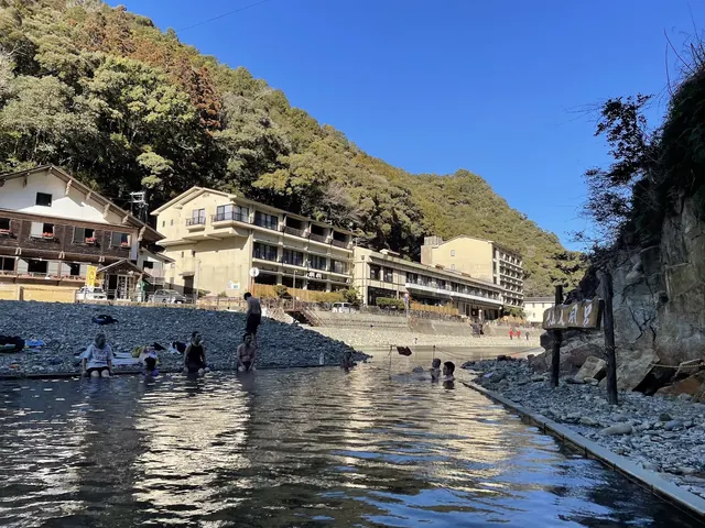 Visitors bathing in Kawayu Onsen Sennin-buro River Bath along a rocky river surrounded by buildings in Tanabe, Japan.