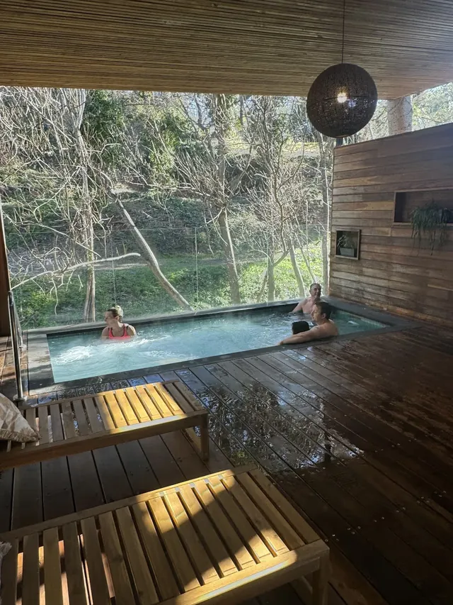Semi-outdoor wooden deck with a rectangular soaking pool where four adults enjoy bathing surrounded by leafless trees in Hepburn Springs Vi.