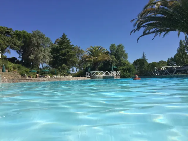 Wide view from water level of Termas de Monfortinho hot spring pool with a swimmer, palm trees, green umbrellas, and wooded hills in Monfor.