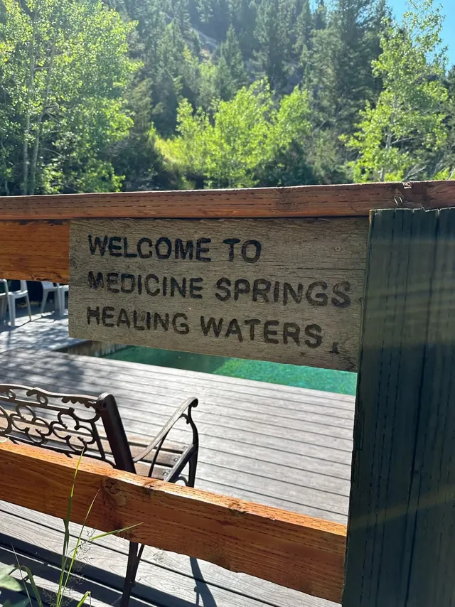 Wooden sign reading Welcome to Medicine Springs Healing Waters with glimpse of a pool, deck, and leafy trees behind on clear sunny day.