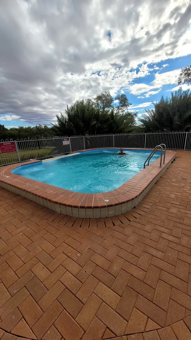 Small heated outdoor spa pool at Bedourie Artesian Spa and Aquatic Centre surrounded by metal fence and green shrubs under cloudy sky.