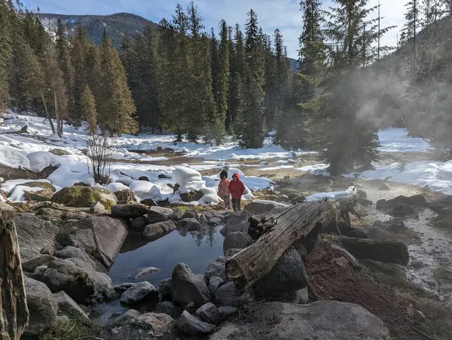 People walking near steaming pools at snow-covered Jerry Johnson Hot Springs with tall pine trees in Kooskia, Idaho, United States.