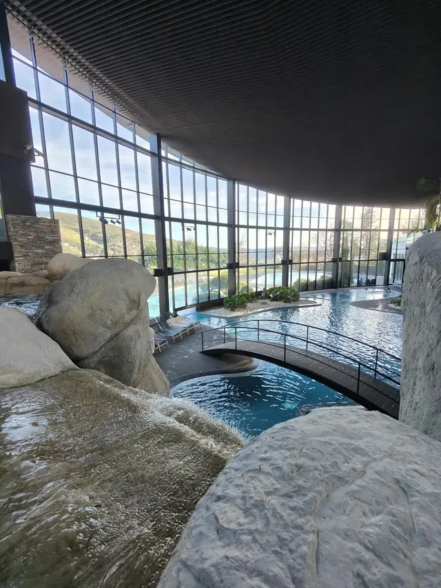 Close-up of rock waterfall flowing into a hot spring pool inside Aquadome with sunlit glass walls and scenic outdoor landscapes in portugal.