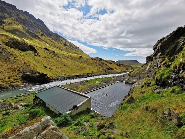 Aerial view of Seljavallalaug hot spring pool in a green valley by a flowing river near Skogar Iceland under a partly cloudy sky.