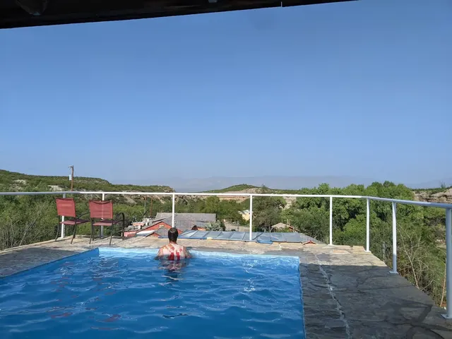 A person soaking in an outdoor hot spring pool with stone decking overlooking green hills near Presidio, Texas at Chinati Hot Springs.