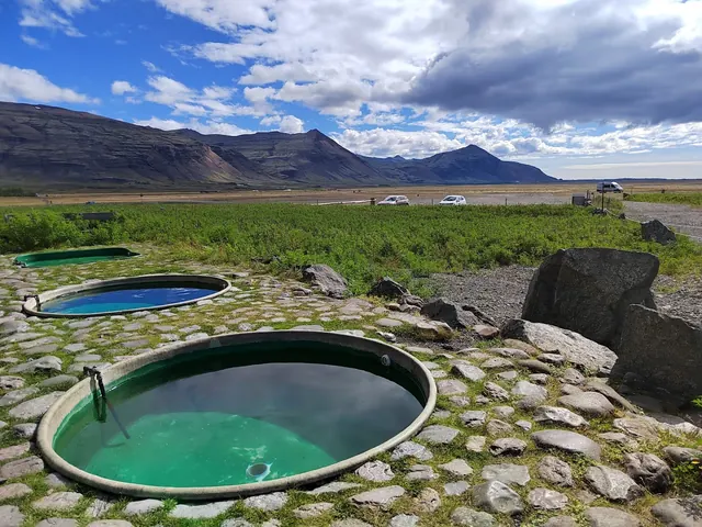 Round mineral pools surrounded by stones steaming slightly with a mountain range near Hofn Iceland in background.