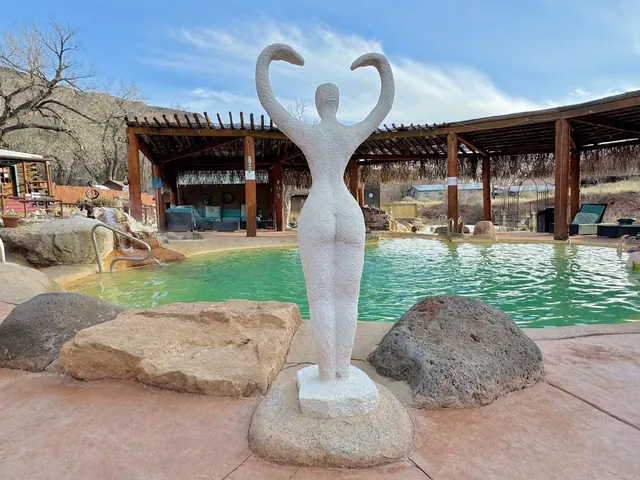 Jemez Hot Springs pool with turquoise water features a white sculpture of a figure raising arms near stone seating and covered lounging are.