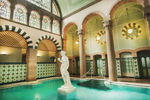 Indoor hot spring pool with turquoise water at Palais Thermal featuring a white statue and green stained glass windows in Bad Wildbad Germa.