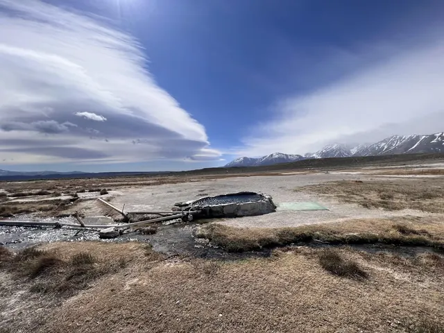 Stone-edged hot spring pool at Hilltop Pulky's Pool with dry grassy field and snow-capped mountains under a blue sky in California.