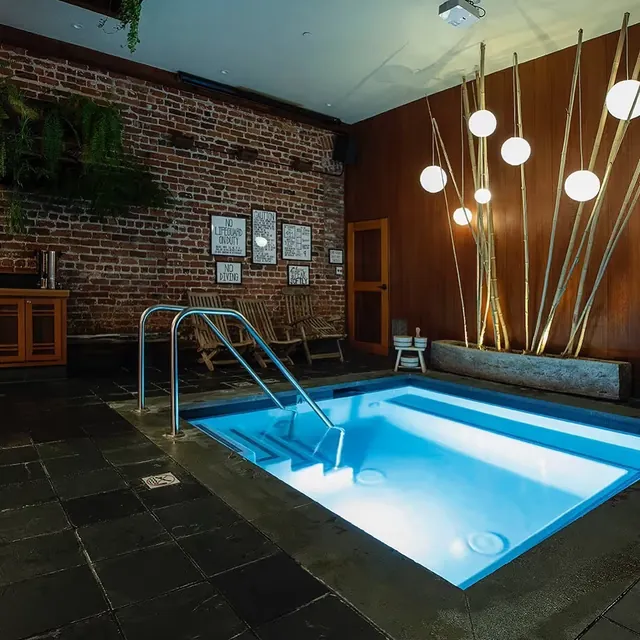 Lit indoor soaking pool at Onsen SF in San Francisco showing metal steps, brick wall, wood seats, and modern hanging lights.