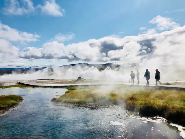 Wooden boardwalk crossing a steaming geothermal hot spring area with hot water pools and hills under a partly cloudy sky at Hveravellir geo.