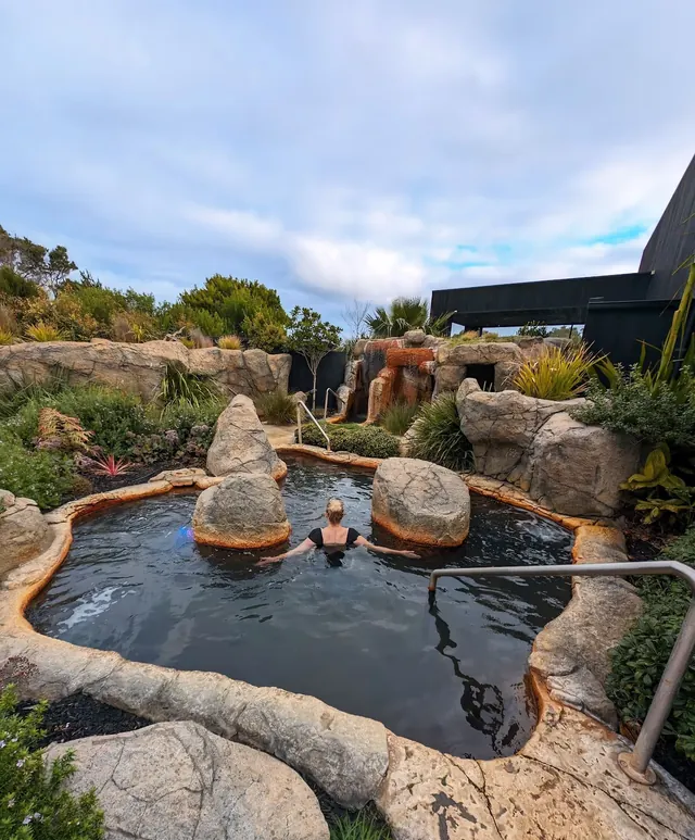 Woman soaking under a rocky cliff waterfall in an outdoor hot spring pool at Deep Blue Hot Springs near Warrnambool Australia.