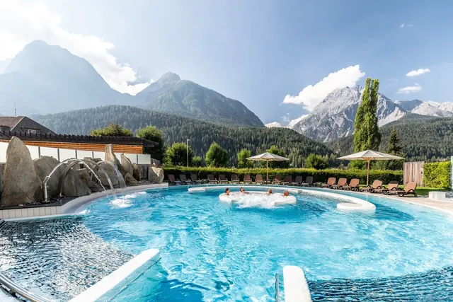 Outdoor hot spring pool at Bogn Engiadina Scuol with water jets, lounge chairs, umbrellas, and a clear sunny mountain landscape.